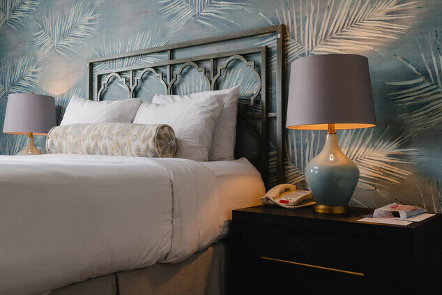 A bed with white bedding and a blue wallpaper with a palm leaf pattern, with two lamps and a nightstand.
